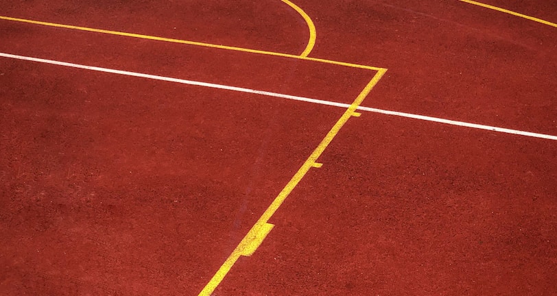 A sports court with a reddish-brown surface and bright yellow and white lines. The lines form geometric shapes, including rectangles and circles, suggesting markings for games like basketball or netball.